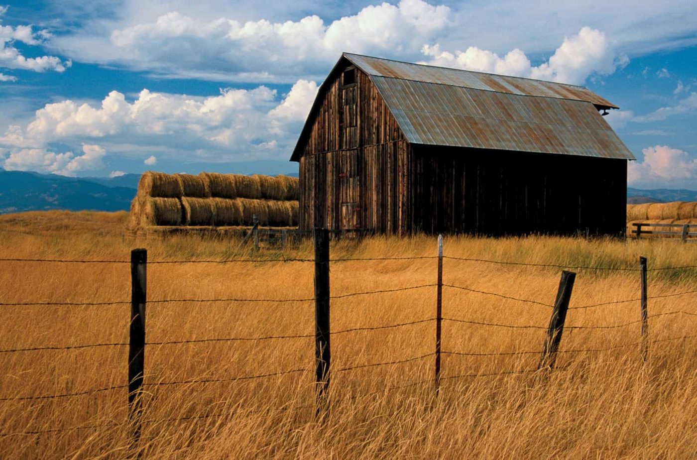 a large wooden barn in the middle of a wheat field next to large rolls of straw