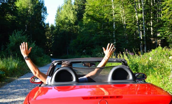 Man driving a red convertible with hands in the air, wife bent down in passengers seat