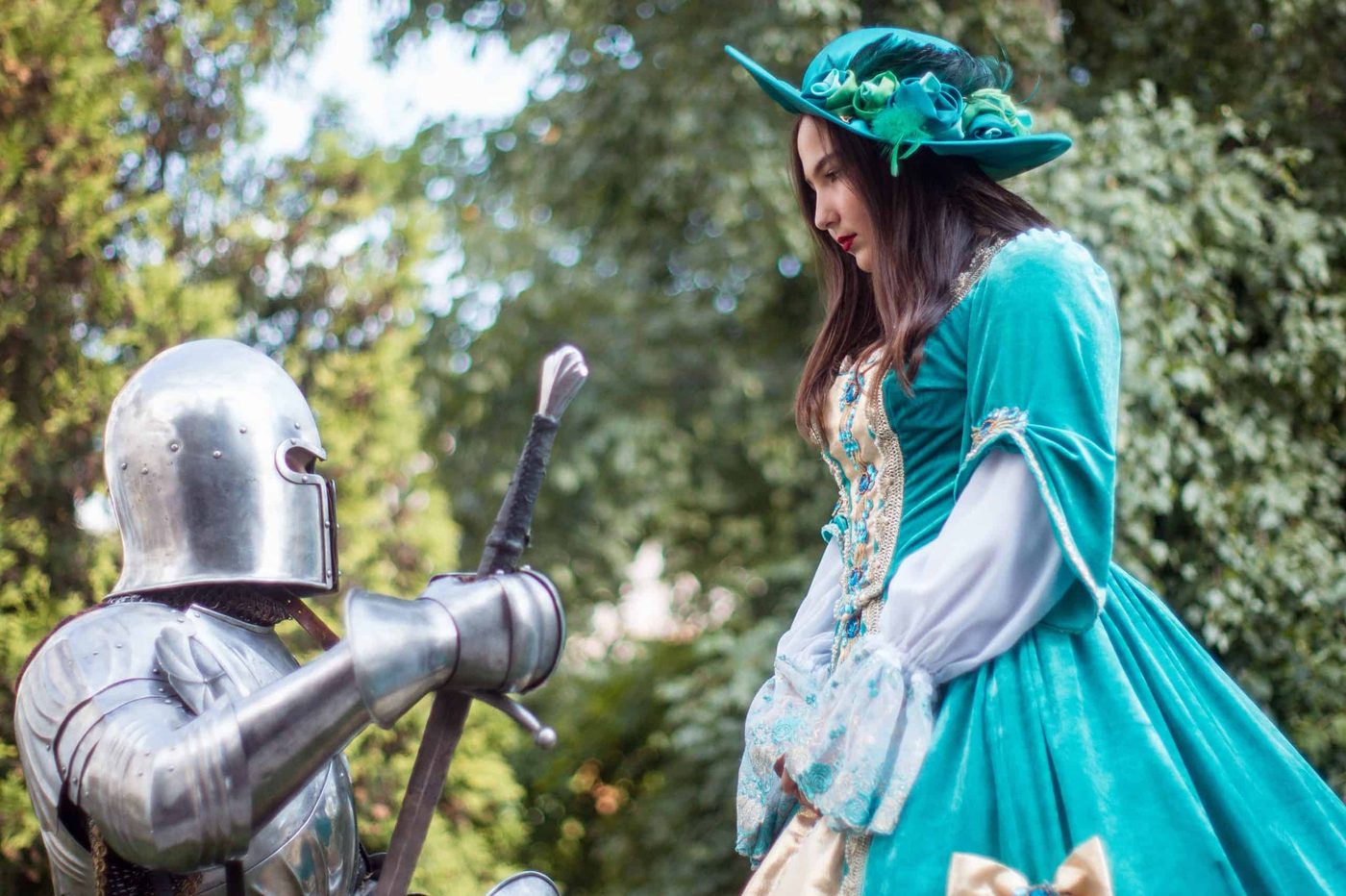 man in steel knight armor holding his sword and kneeling infront of a woman in dress and flowery hat