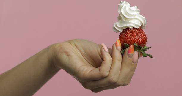Woman hand holds strawberry with whipped cream on top of the berry. Pink background
