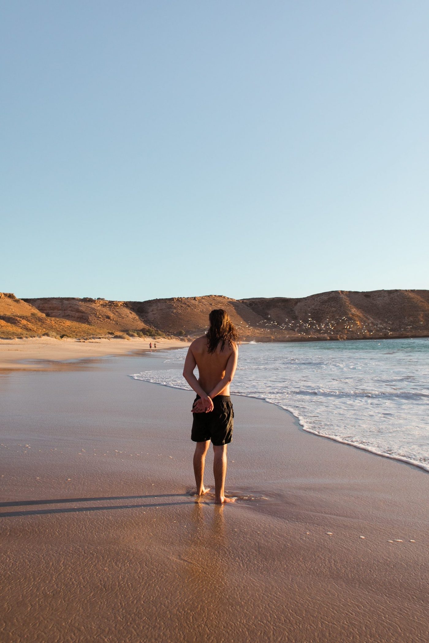 Man standing on the water line at the beach in bathing suit and holding his hands behind his back
