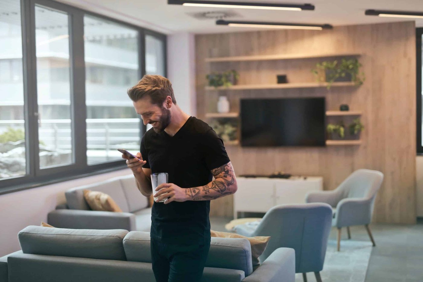 Man standing in the office smiling while looking at his phone and holding a drinking glass