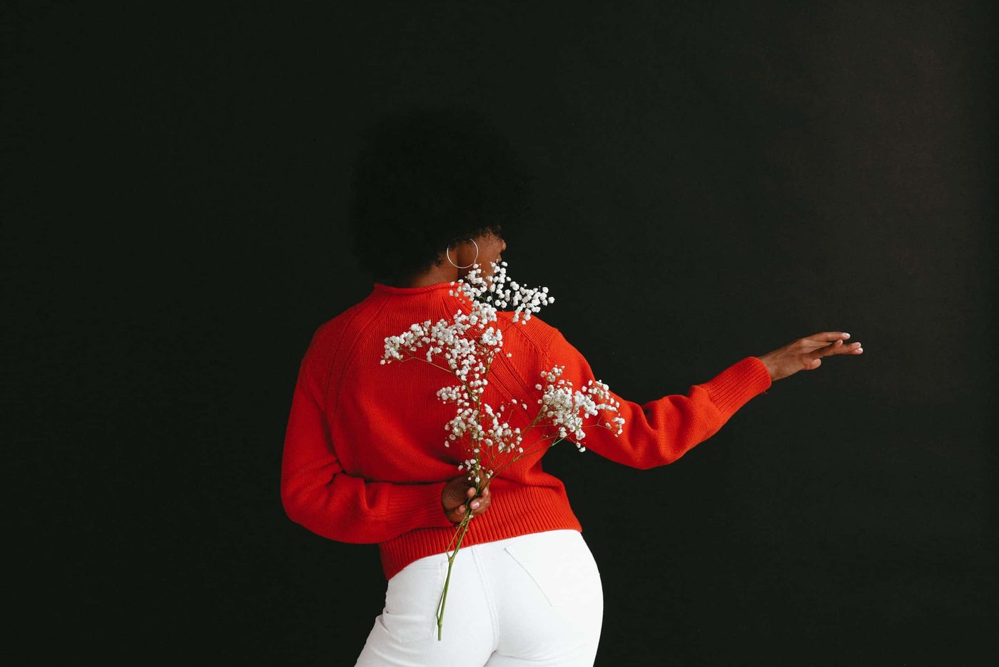 woman with back turned wearing red sweater, holding flowers behind her back and gesturing away with the other hand