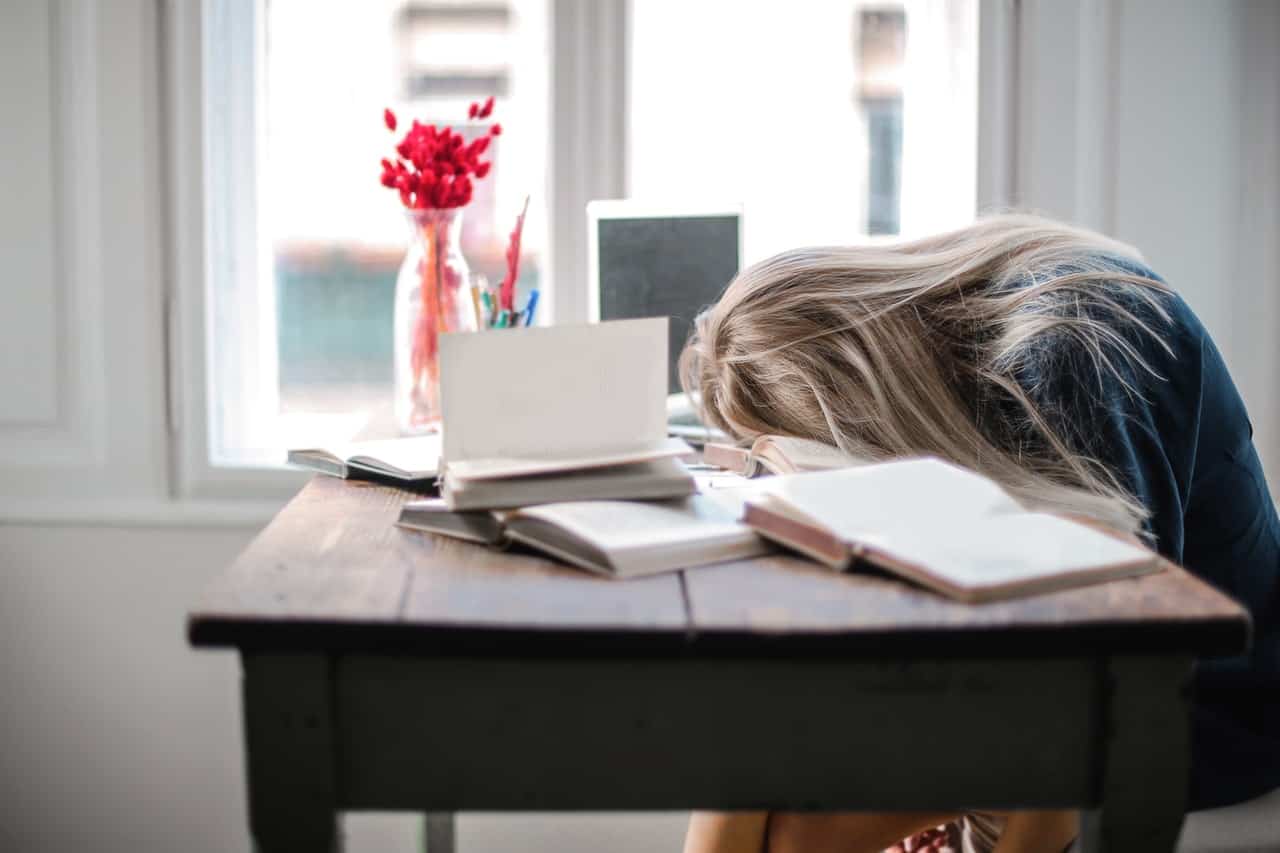 Frustrated and tired woman resting her head on work desk, computer screen and books are lying around