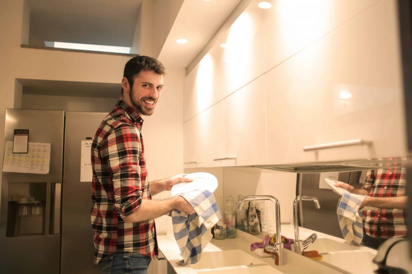Man washing dishes at the kitchen sink while his partner photographs him