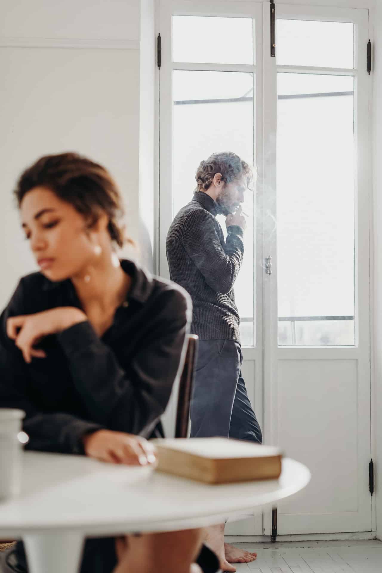 Couple having a misunderstanding, woman at the table looking upset and an upset man smoking by the window
