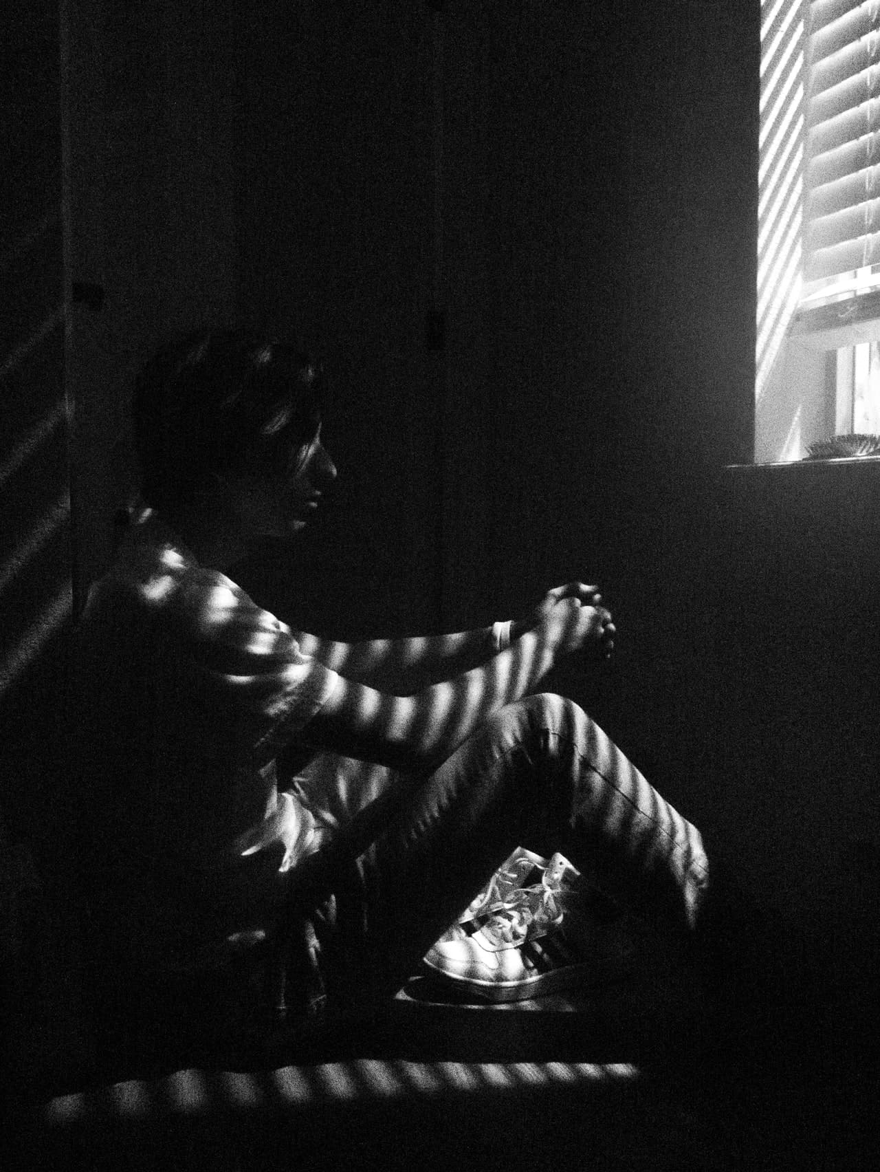 man looking despaired sitting on his bed, hugging his legs and looking out the window