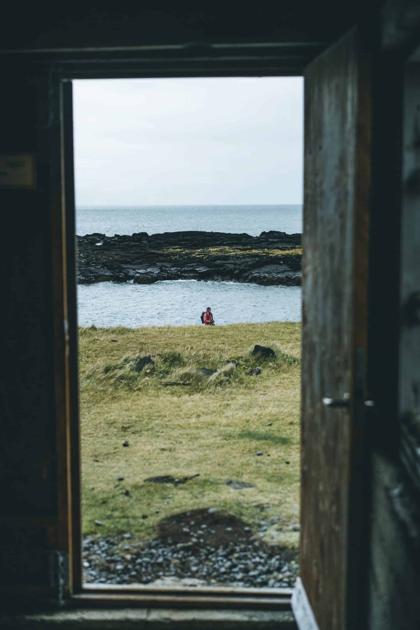 Open door leading to a grassy lakeside beach with a person sitting by the water