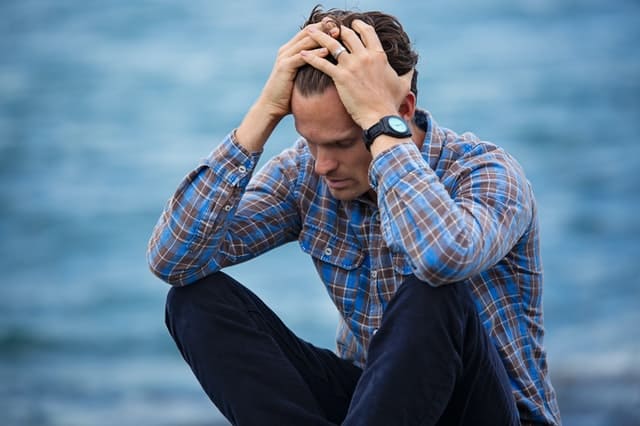 stressed guy sitting by the ocean holding his head with both his hands and pulling his hair back