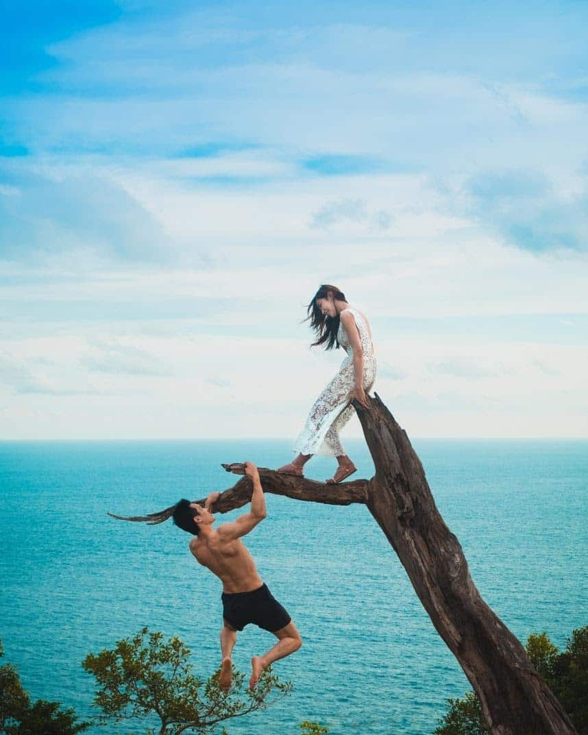 woman sitting on a dead tree at the beach, man hanging off a branch