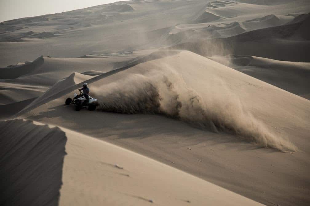 quadbike driving over a sand dune with a cloud of dust behind him