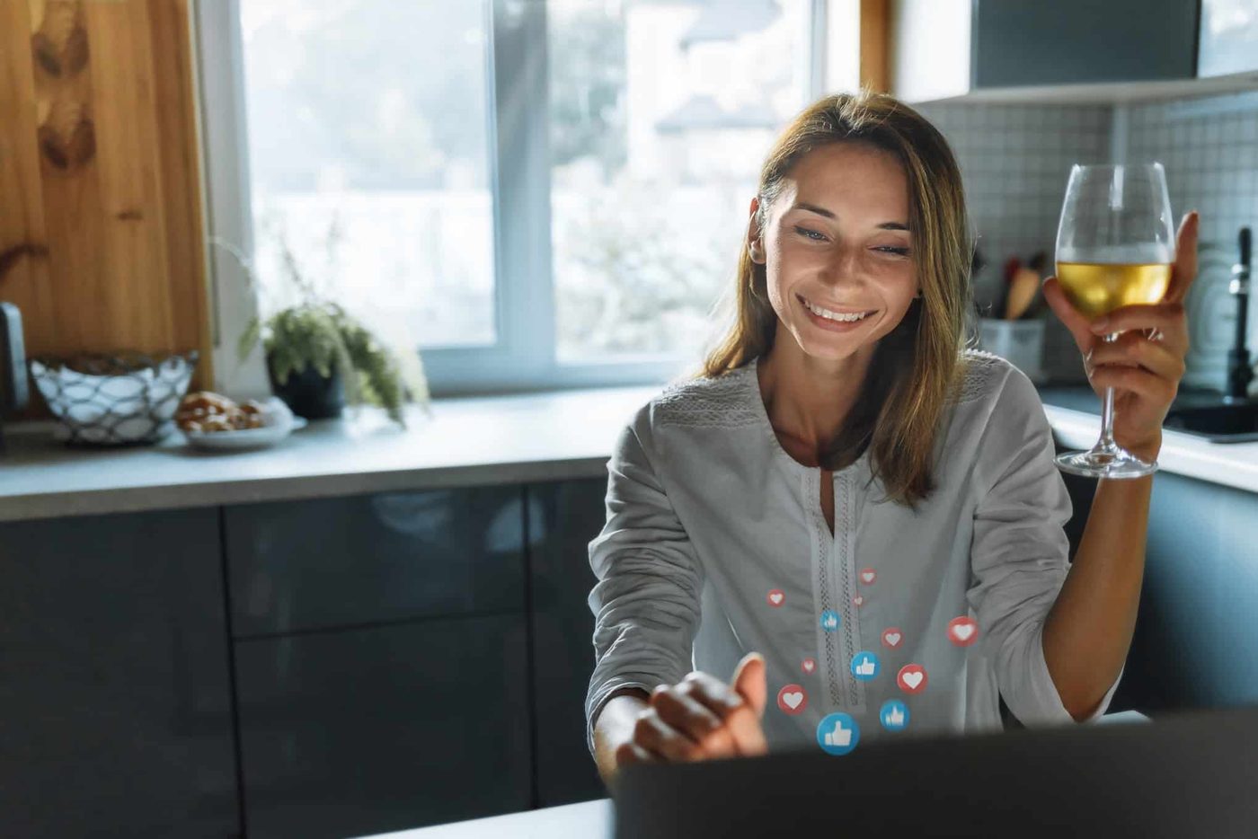 Happy young woman with glass wine talking through social network sitting at home in the kitchen